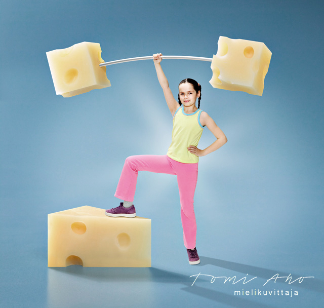 Nuori voimatyttö kannattelee levypainotankoa, jossa painoina on juustokuutiot. Mielikuvittaja Tomi Aho