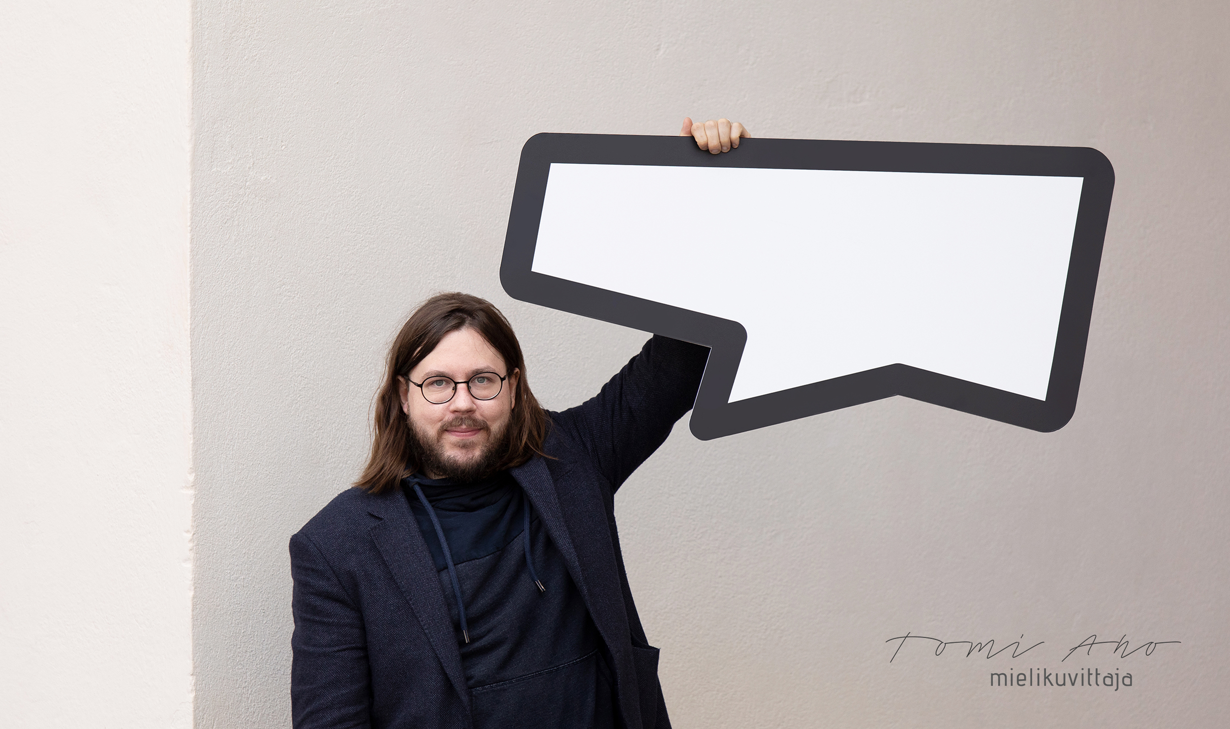 Pitkähiuksinen mies pitää puhekuplaa kädellään Tomi Ahon valokuvaamassa markkinointikuvassa.