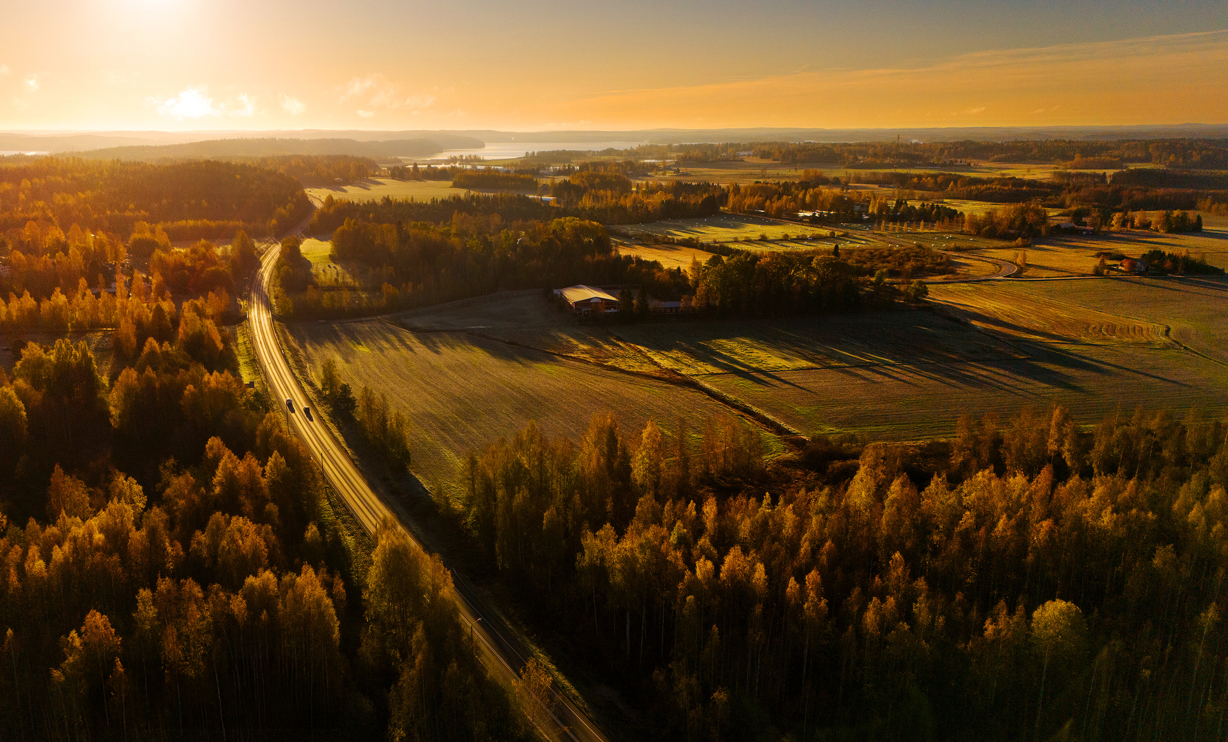 Dronekuva maisemasta, jossa aurinko laskee ja alla näkyy pitkä tie, jossa autot pieniä, Mielikuvittaja Tomi Aho.